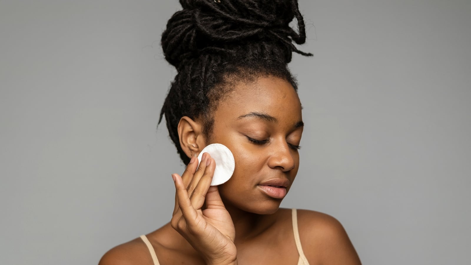 Gentle young ethnic female with Afro braid bun and closed eyes purifying face skin with cotton disk on gray background.
